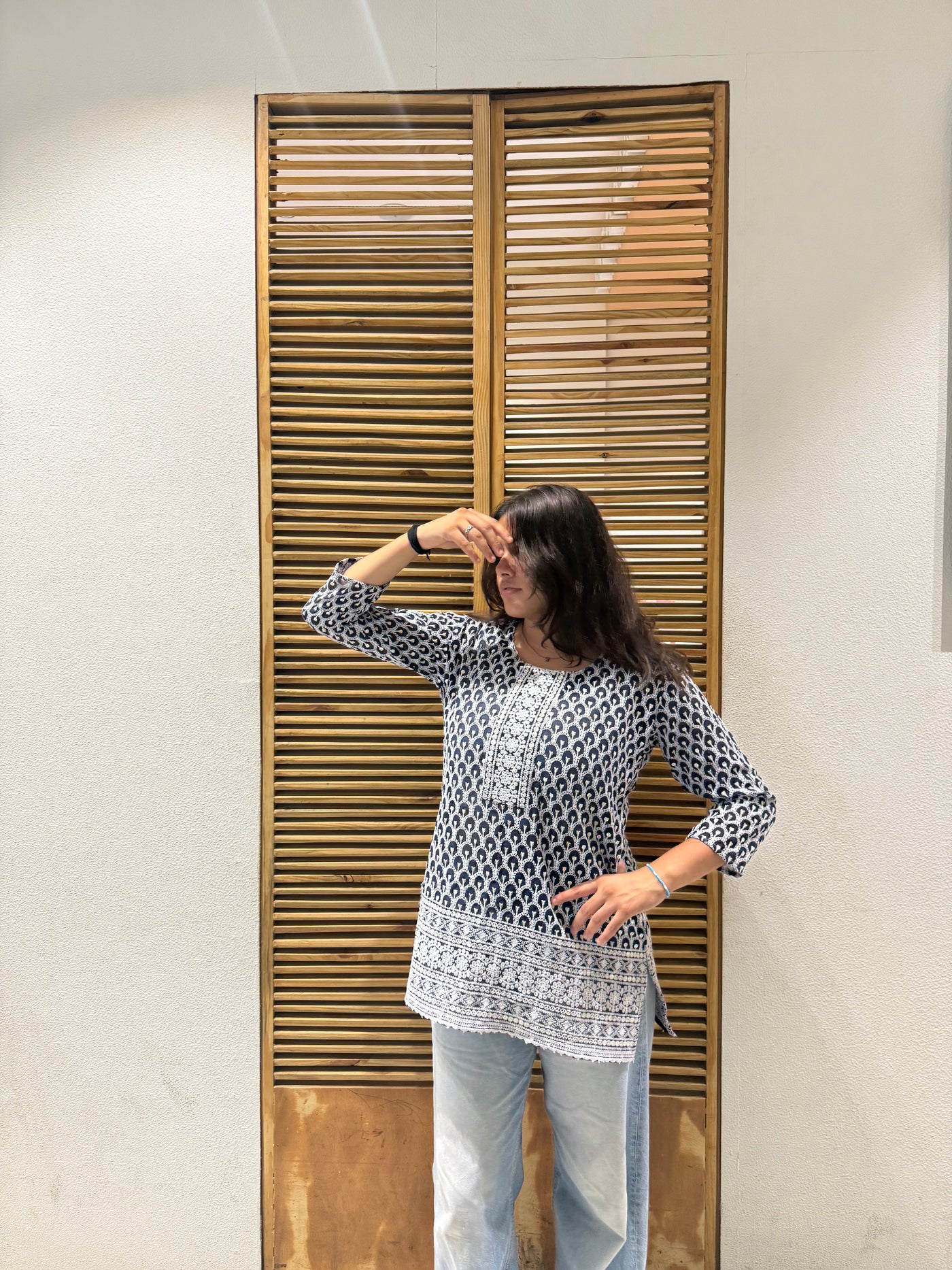 Woman standing in front of a wooden door with slats, wearing a patterned top and light blue jeans.