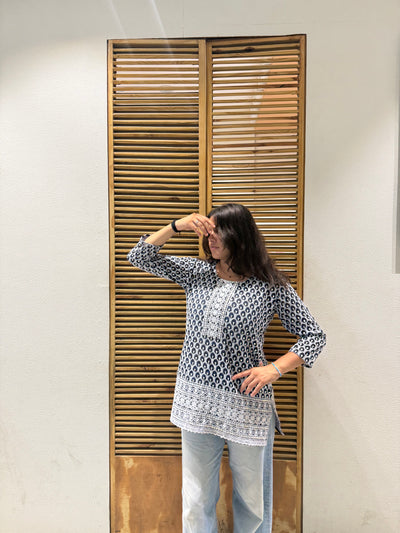 Woman standing in front of a wooden door with slats, wearing a patterned top and light blue jeans.