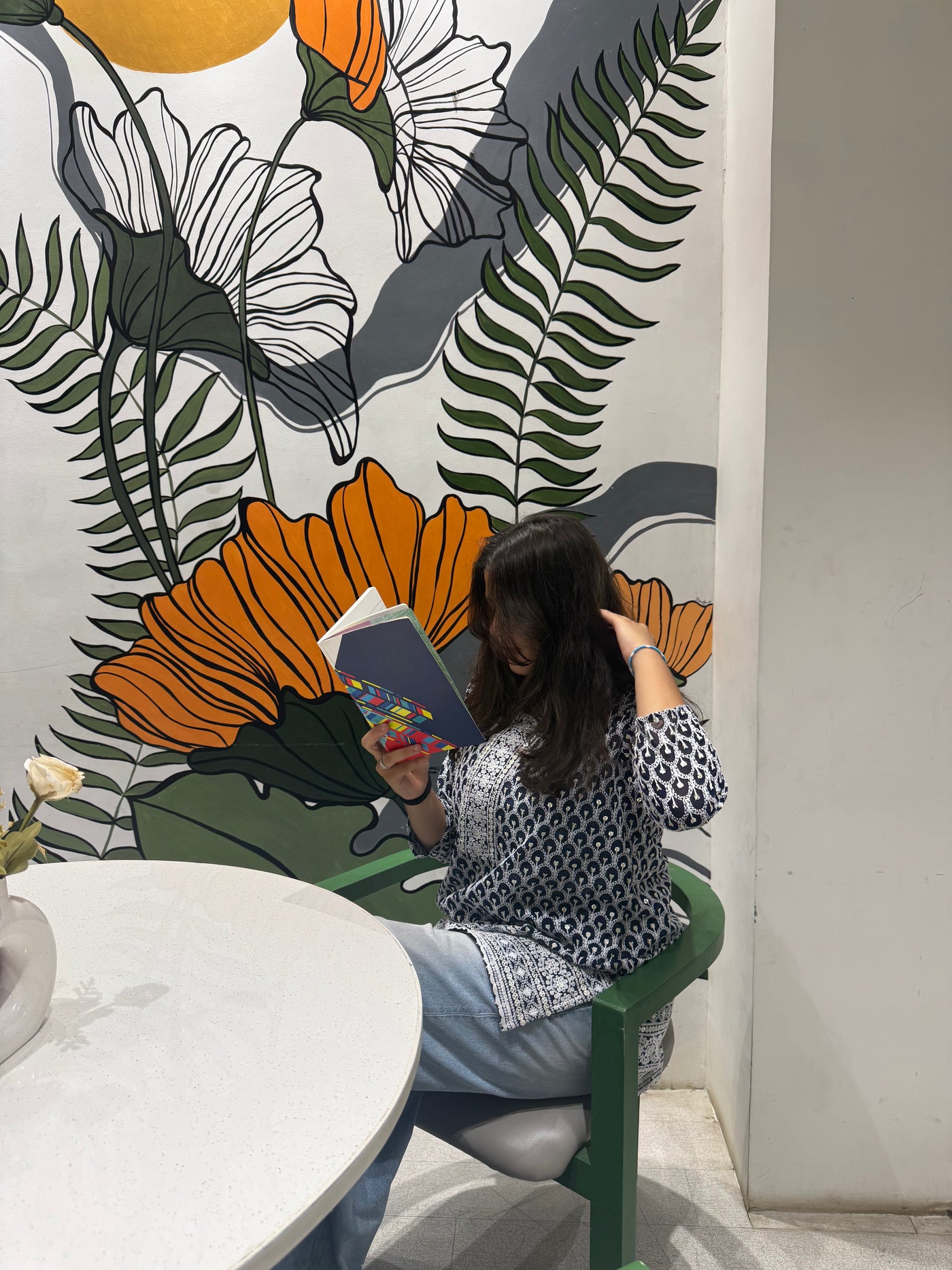 Person sitting on a green stool reading a book against a colorful floral mural.