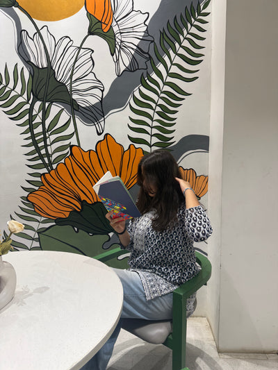 Person sitting on a green stool reading a book against a colorful floral mural.